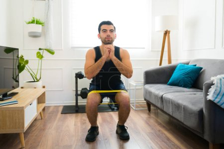 Téléchargez les photos : Fitness homme sportif dans sa vingtaine faire des exercices de squat en utilisant une bande de résistance tout en travaillant à la maison - en image libre de droit