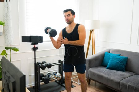 Téléchargez les photos : Forte forme homme soulevant haltères poids tout en filmant une vidéo de fitness pour les médias sociaux dans son salon - en image libre de droit