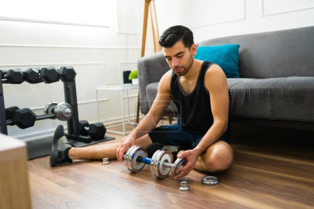 Photo for Sporty fit man in his 20s putting weights in his dumbbells to start lifting during his home workout - Royalty Free Image