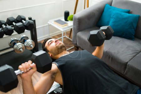 Foto de High angle of an hispanic man at his home gym doing bench press with dumbbell weights - Imagen libre de derechos
