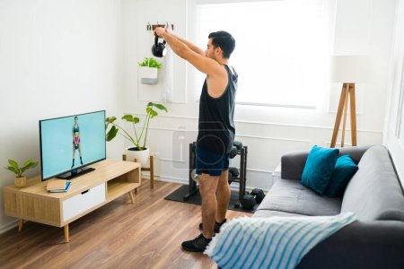 Téléchargez les photos : Jeune homme en bonne santé s'entraînant à soulever un poids de kettlebell tout en regardant une vidéo d'entraînement à domicile dans le salon - en image libre de droit