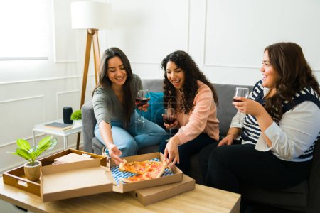 Foto de Excited female best friends laughing eating pizza and drinking wine together at home while having fun together - Imagen libre de derechos
