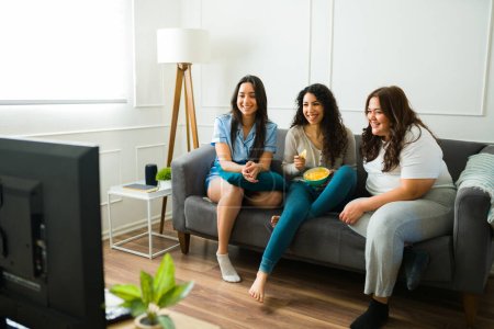 Téléchargez les photos : Happy female friends laughing and eating snacks while relaxing together watching a movie in the living room during a sleepover - en image libre de droit
