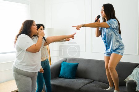 Photo for Hispanic young women in pajamas having fun playing karaoke and singing with music during their sleepover at home - Royalty Free Image