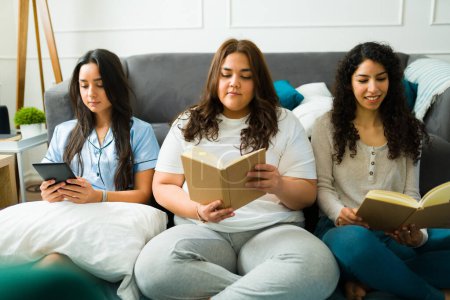 Foto de Beautiful female friends in pajamas relaxing reading a book together in pajamas while having a sleepover at home - Imagen libre de derechos