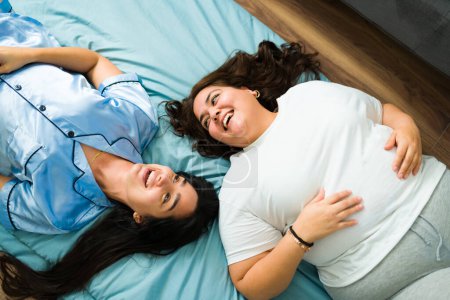 Foto de Top view of female best friends laughing and joking while having fun relaxing in bed at their sleepover - Imagen libre de derechos