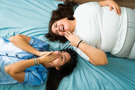 Foto de Happy latin women and best friends laughing together while lying in bed in their pajamas - Imagen libre de derechos
