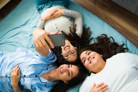 Foto de High angle of three young female friends taking a selfie with their phone while relaxing in pajamas during a sleepover - Imagen libre de derechos