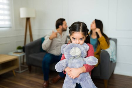 Entzückendes kleines Mädchen, das einen Teddybär umarmt und ängstlich und traurig schaut, während es ihren Eltern zuhört, wie sie im Wohnzimmer kämpfen