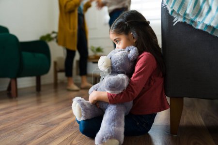 Photo for Sad young kid sitting alone hugging a teddy bear while looking scared about mom and dad screaming and fighting at home - Royalty Free Image