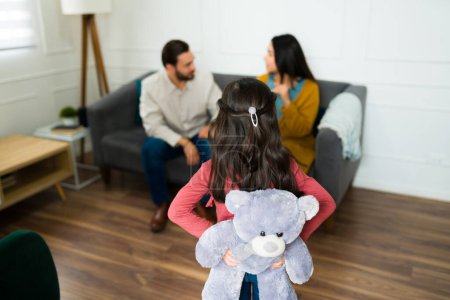Kleines Kind von hinten gesehen, wie es einen Teddybär hält, während es seine wütenden Eltern ansieht, die über das Sorgerecht nach der Scheidung sprechen