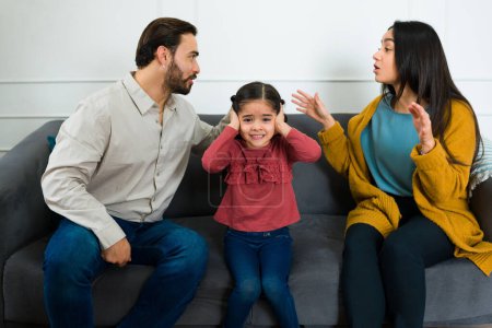 Niña triste hispana cubriéndose los oídos mientras escucha a su madre y su padre gritando tener problemas de matrimonio en casa