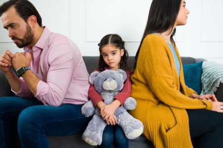 Niedliches trauriges kleines Kind mit einem Teddybär, der Blickkontakt herstellt, während es nach einem üblen Kampf inmitten seiner wütenden Eltern sitzt