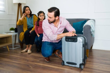 Trauriger Ehemann und Vater mit einem Koffer, der von zu Hause wegzieht und seine verärgerte, wegen Scheidung auseinandergerissene Familie betrachtet