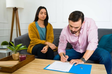 Photo for Caucasian angry man signing the divorce papers with a sad hispanic woman sitting on the sofa at home - Royalty Free Image