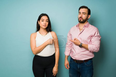 Photo for Attractive annoyed couple pointing and blaming each other after fighting or arguing in front of a studio background - Royalty Free Image