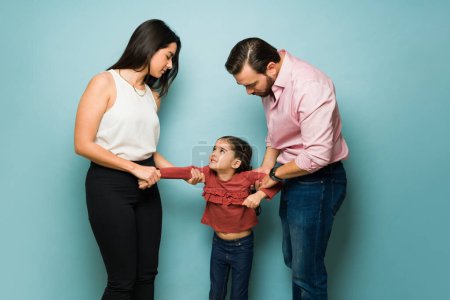 Photo for Divorced couple fighting about child custody and trying to take their scared young daughter in front of a blue background - Royalty Free Image