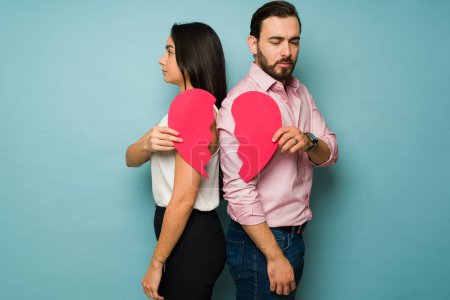 Photo for Angry attractive couple feeling heartbroken turning their backs holding a red broken heart after breaking up - Royalty Free Image