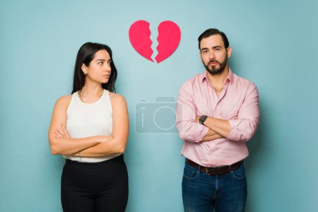Upset latin woman looking annoyed at her partner while feeling heartbroken against a studio background with a broken heart
