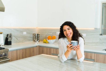 Photo for Excited beautiful woman in her beautiful white kitchen laughing and looking happy while enjoying drinking a cup of coffee - Royalty Free Image