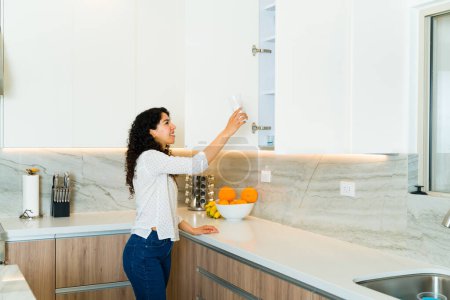 Foto de Joven hispana alegre buscando una taza o plato en su hermoso armario blanco en una cocina de lujo - Imagen libre de derechos