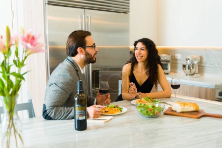 Téléchargez les photos : Joyeux couple souriant mangeant une salade lors d'un dîner assis à leur îlot de cuisine en granit regardant heureux - en image libre de droit