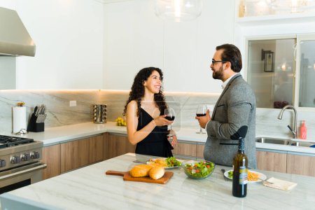 Photo for Attractive couple laughing and having fun while drinking wine in a luxury kitchen before an elegant date at home - Royalty Free Image