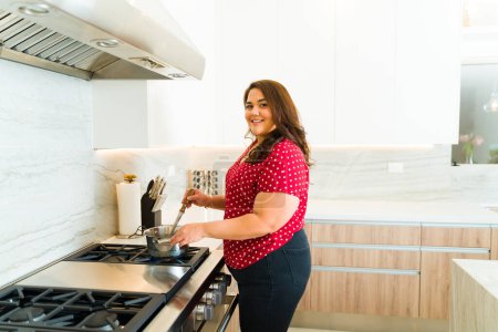 Foto de Feliz mujer latina gorda sonriendo haciendo contacto visual mientras cocina el almuerzo usando la estufa en su hermosa cocina de lujo - Imagen libre de derechos