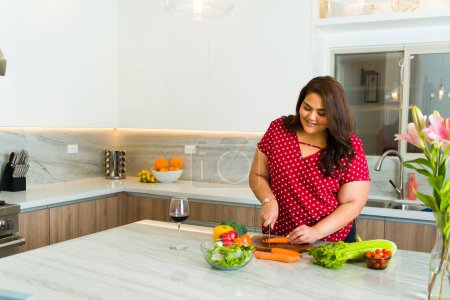 Foto de Mujer obesa hispana sonriendo mientras corta verduras para cocinar la cena en su cocina de lujo de granito - Imagen libre de derechos