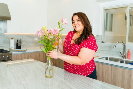 Téléchargez les photos : Attrayant graisse jeune femme souriant faire contact visuel à sa grande cuisine blanche de granit tout en faisant un arrangement de fleurs - en image libre de droit