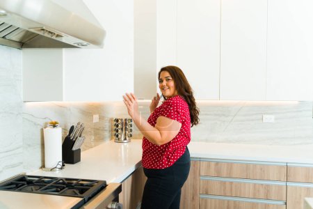 Téléchargez les photos : Souriant hispanique femme obèse souriant regardant heureux dans sa belle cuisine de luxe tout en ouvrant le placard en granit - en image libre de droit