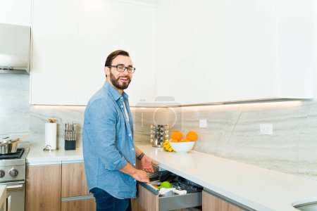 Téléchargez les photos : Joyeux homme caucasien attrayant souriant ouvrant le tiroir de comptoir en granit blanc dans sa cuisine de luxe - en image libre de droit