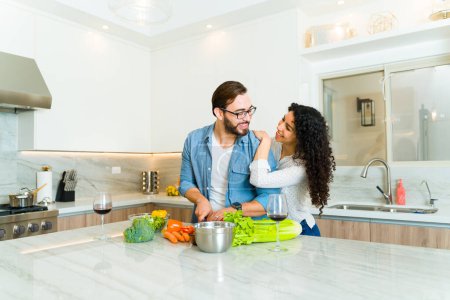 Téléchargez les photos : Couple romantique souriant regardant en amour tout en embrassant la cuisine d'un dîner sain ensemble dans leur cuisine de luxe - en image libre de droit