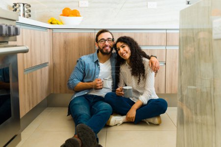 Foto de Alegre hombre caucásico y mujer hispana abrazándose mientras sonríen tomando café juntos en el piso de la cocina - Imagen libre de derechos