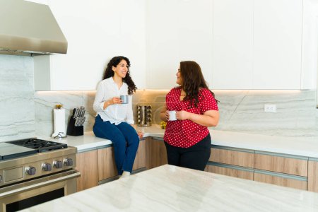 Photo for Cheerful young women and best friends relaxing while chatting and drinking coffee together in a luxury kitchen - Royalty Free Image