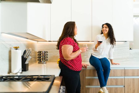 Photo for Smiling latin girlfriends enjoying drinking coffee together in a beautiful white kitchen and looking happy and relaxed - Royalty Free Image