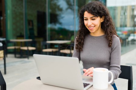 Foto de Hermosa mujer feliz con el pelo rizado sonriendo en la cafetería mientras que hace el trabajo remoto y escribiendo en el ordenador portátil - Imagen libre de derechos