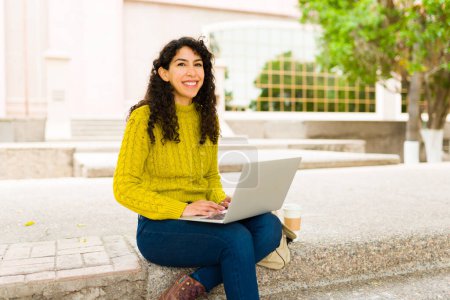 Téléchargez les photos : Belle femme heureuse avec les cheveux bouclés souriant et travaillant sur son ordinateur portable à l'extérieur faisant du travail à distance - en image libre de droit