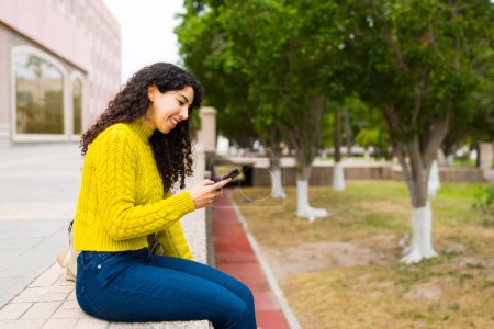 Téléchargez les photos : Vue latérale d'une belle femme heureuse assise dans le parc à l'extérieur et souriante tout en envoyant des SMS à des amis sur le smartphone - en image libre de droit