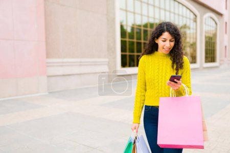 Foto de Alegre mujer caucásica divertirse mensajes de texto después de ir de compras y llevar un montón de bolsas fuera del centro comercial - Imagen libre de derechos