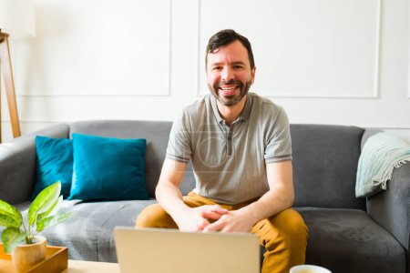 Foto de Atractivo hombre caucásico mirando feliz sonriendo mientras usa su computadora portátil en la sala de estar y trabajando desde casa - Imagen libre de derechos