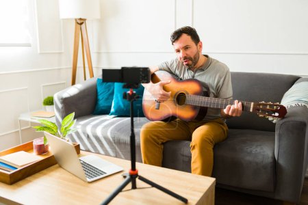 Foto de Talentoso hombre artístico filmando un video musical con una cámara mientras vlogging tocando la guitarra en su casa - Imagen libre de derechos