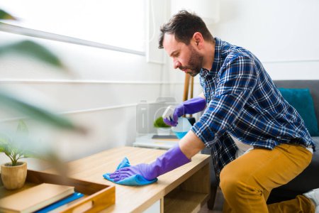 Photo for Handsome caucasian man wearing gloves and cleaning the coffee table of the living room while doing chores at home - Royalty Free Image