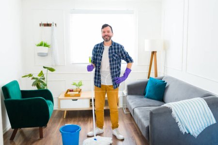 Foto de Retrato de un hombre caucásico feliz sonriendo mirando a la cámara mientras se prepara para limpiar su sala de estar usando una fregona - Imagen libre de derechos