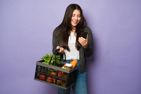 Foto de Feliz mujer caucásica mensajes de texto en su teléfono inteligente mientras usa una cesta para hacer sus compras de comestibles - Imagen libre de derechos