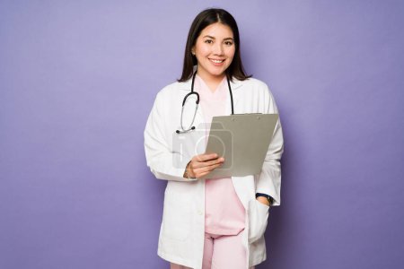 Téléchargez les photos : Magnifique femme caucasienne souriant à l'aide d'un manteau blanc regardant la caméra tenant un presse-papiers pour un patient - en image libre de droit