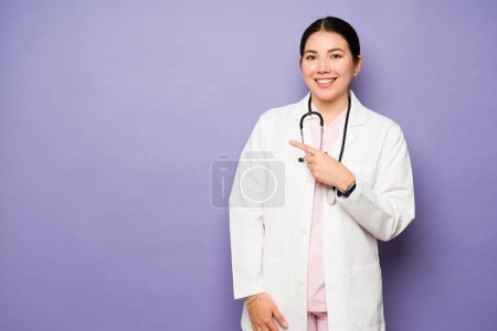 Téléchargez les photos : Portrait d'une femme médecin souriante portant un manteau blanc et un stéthoscope pointant vers la copie de l'espace sur un fond de studio - en image libre de droit