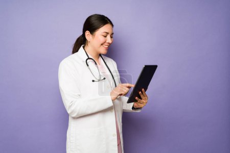 Foto de Perfil de una mujer feliz médico usando una tableta y sonriendo mientras trabajaba en el hospital aislado sobre un fondo púrpura - Imagen libre de derechos