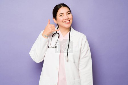 Téléchargez les photos : Magnifique médecin caucasien féminin portant un manteau blanc faisant un geste d'appel téléphonique et établissant un contact visuel - en image libre de droit