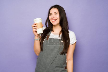 Photo for Cheerful young woman with an apron working as a female barista smiling while serving a coffee to go at the cafe - Royalty Free Image